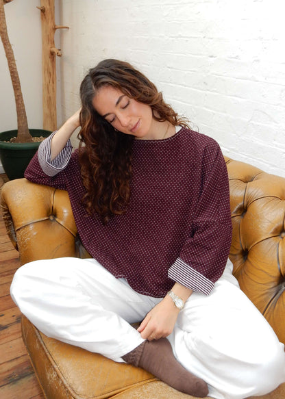 A woman sits cross-legged on a tan leather sofa in a bright room, wearing a burgundy long-sleeve knit with a small white pattern, white jeans, and brown socks.