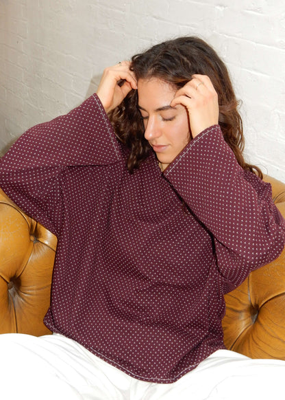 A woman with long brown hair sits on a tan leather couch, wearing a loose-fitting burgundy knit with a small white pattern and white jeans.