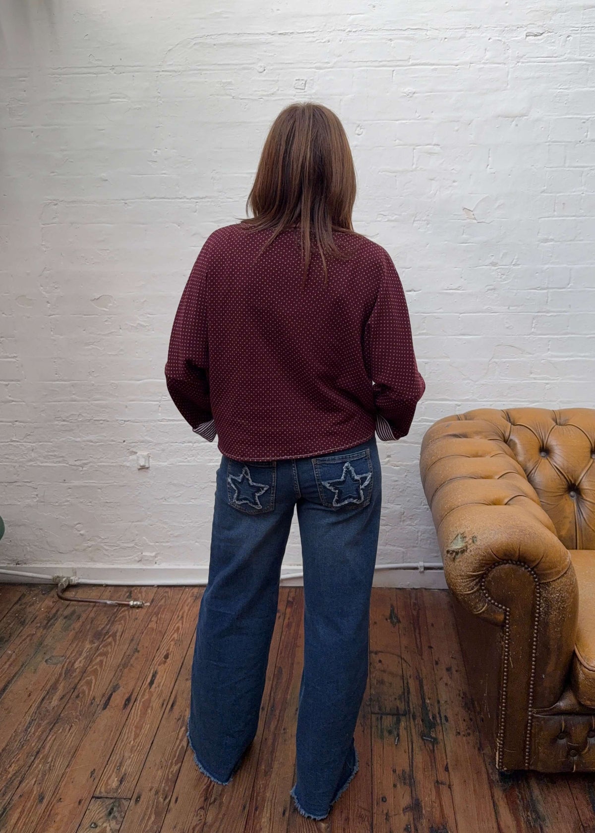 A woman photographed from behind wearing a burgundy long-sleeve knit top and blue jeans with star-shaped patch pockets, standing in a room with a white brick wall, wooden floor, and a tan leather sofa.