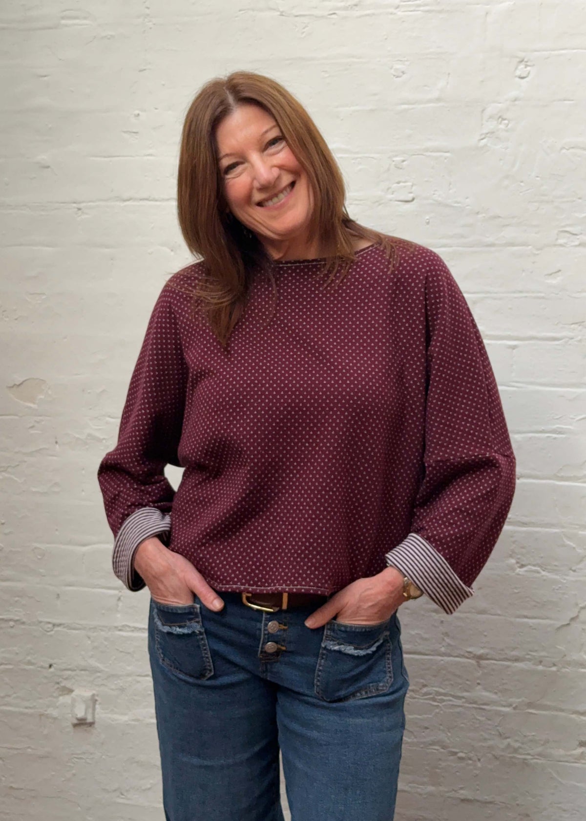 A woman with shoulder-length brown hair stands against a white painted brick wall, wearing a burgundy knit with striped cuffs and blue jeans.
