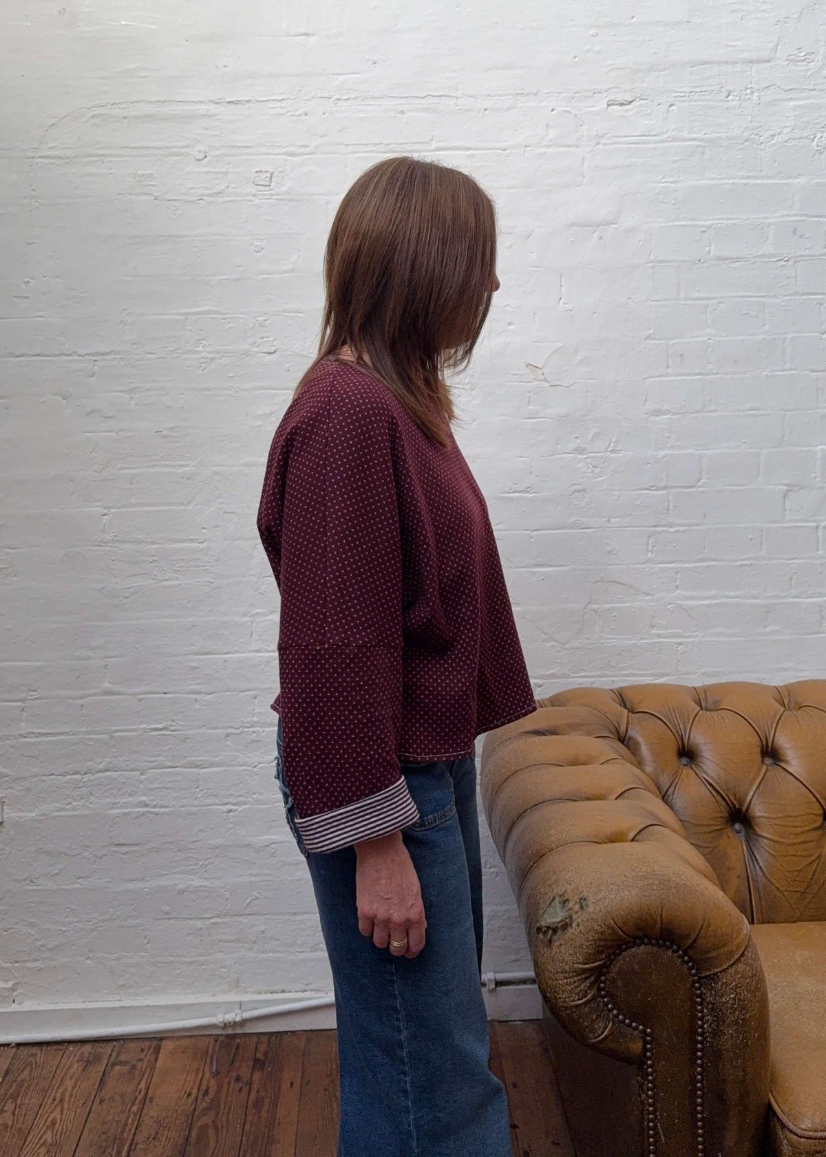 A woman stands in a side profile wearing a burgundy knit with a striped cuff and blue jeans, standing in a room with a white brick wall, wooden floor, and a tan leather sofa.