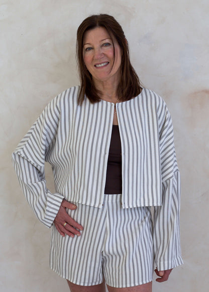 A woman modeling a matching two-piece set consisting of a long-sleeve striped jacket and coordinating striped shorts, worn over a dark brown top. She is standing against a textured, light beige plaster wall.