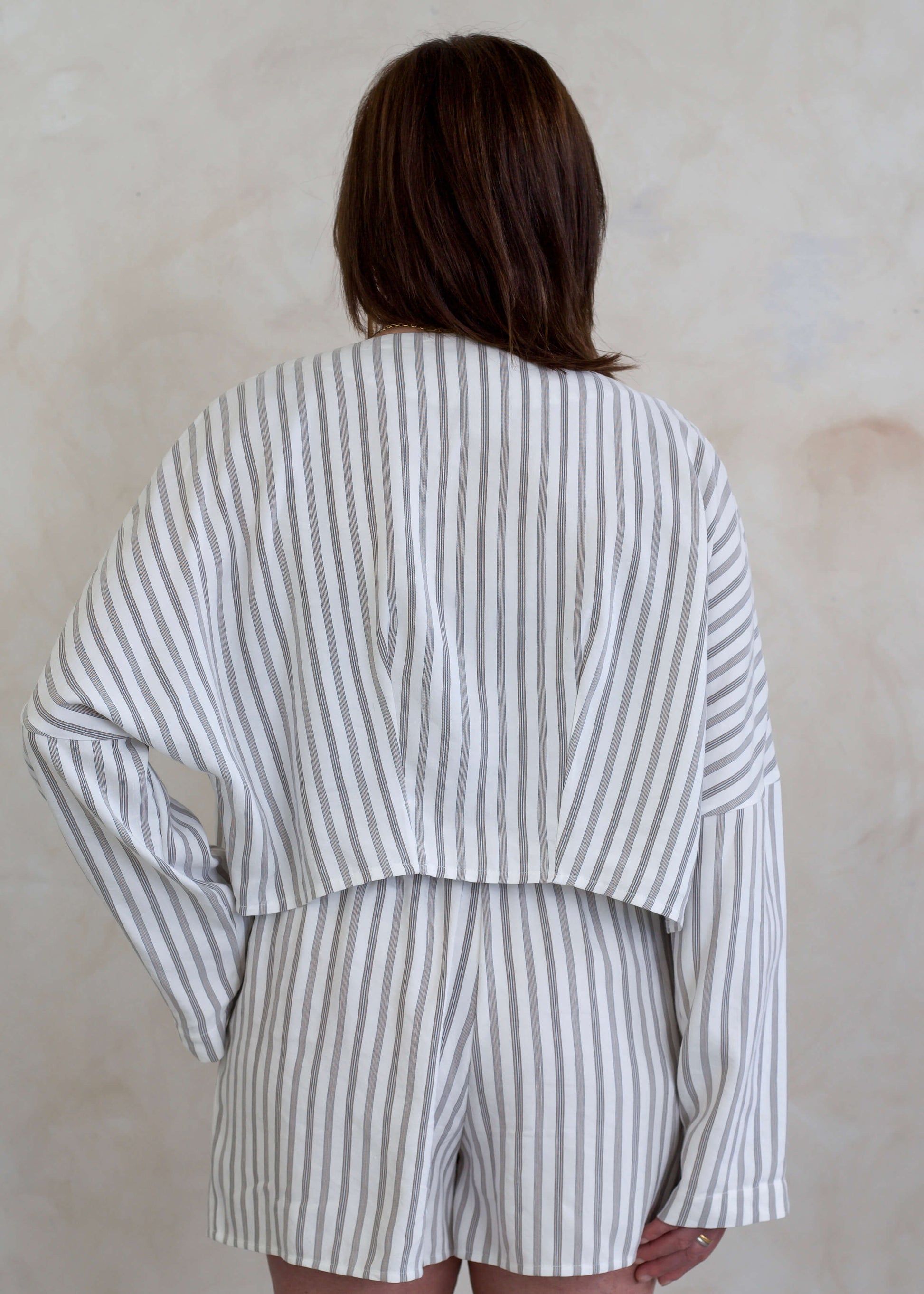 A person shown from the back wearing a matching striped long-sleeve jacket and shorts set in a soft off-white and brown striped pattern, set against a textured beige wall.